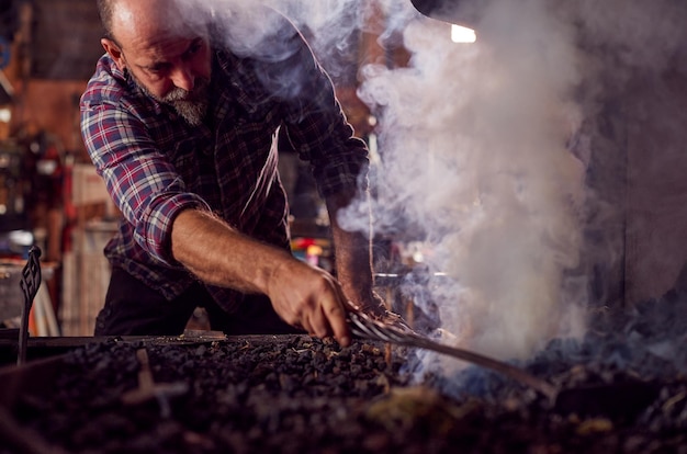 Male Blacksmith Raking Coals Fuel To Start Blaze In Forge