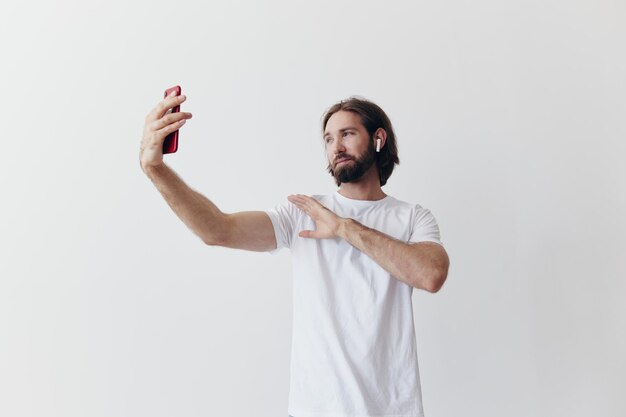 A male blogger videotaping himself on his phone and chatting with people online with a smile in a white Tshirt against a white wall High quality photo