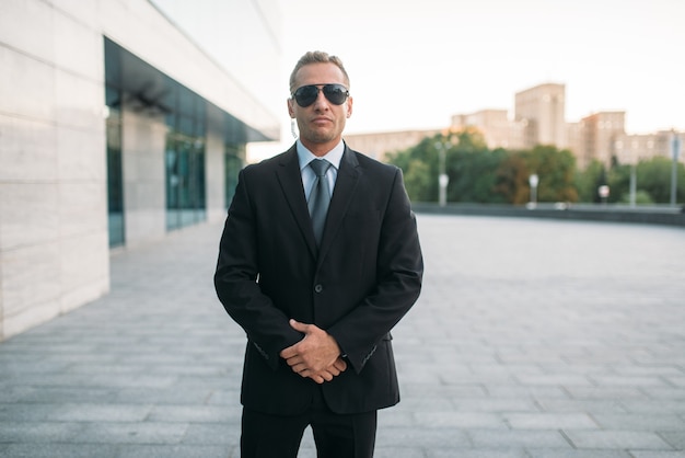 Male bodyguard in suit, security earpiece and sunglasses outdoors.