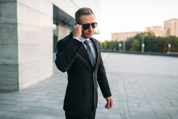 Male bodyguard in suit and sunglasses talking by security earpiece  outdoors. Professional guarding is a risky profession