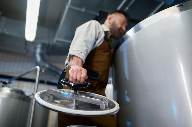 Мужчина-пивовар в фартуке заглядывает внутрь пивного бака, чтобы контролировать процесс пивоварения