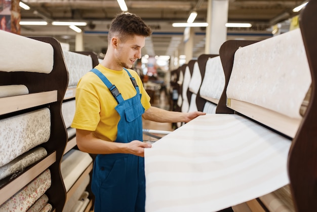 Male builder choosing wallpapers in hardware store. Constructor in uniform look at the goods in diy shop