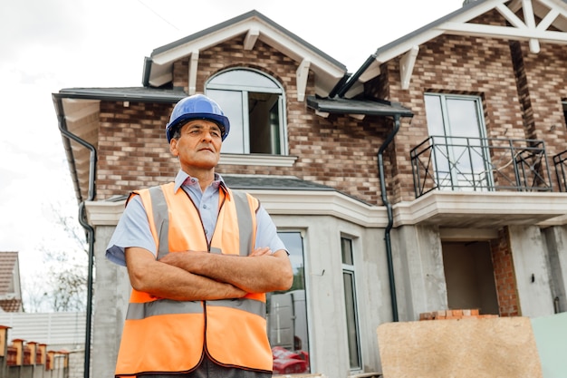 Male builder contractor stands with his crossed hands smiling and looking away wearing a safety helmet and vest with a background construction unfinished home