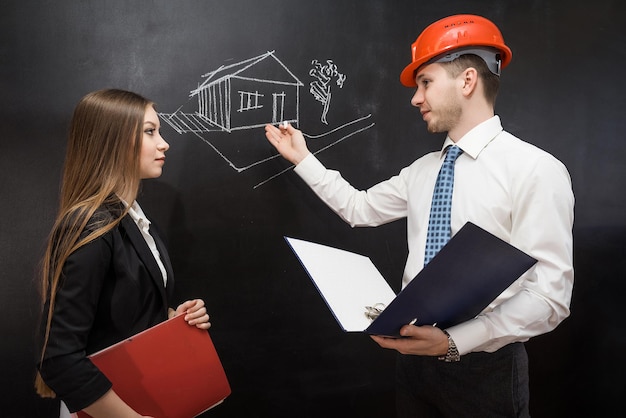 Male builder demonstrate house draw on chalkboard to his client with folder in hands