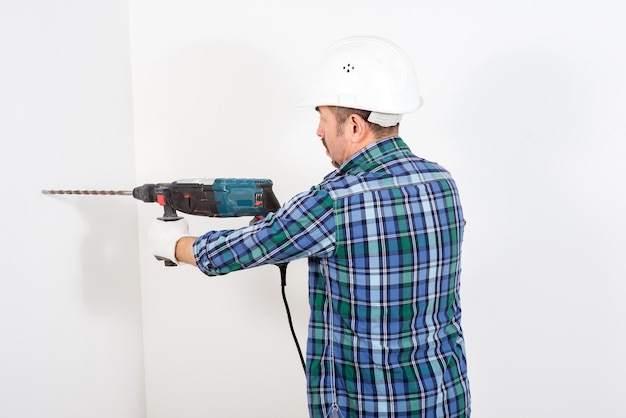 A male builder in a hardhat with a puncher drills a white wall