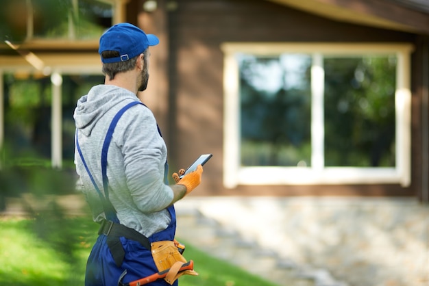 male builder in uniform using smartphone  while working at cottage construction site