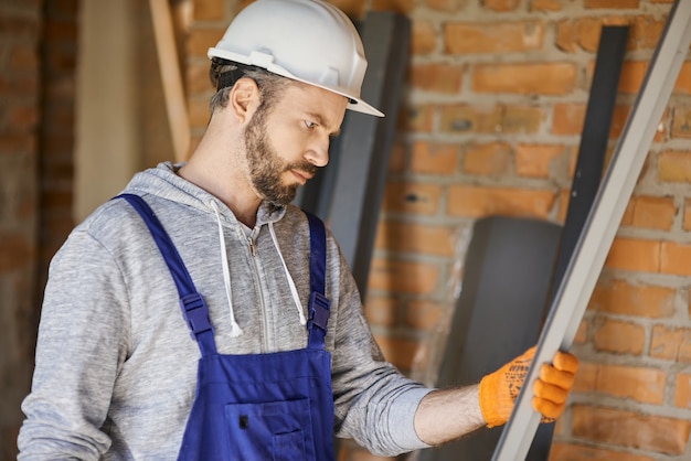 Строитель-мужчина в комбинезоне и каске держит металлический стержень для гипсокартона на внутренней строительной площадке