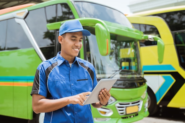 A male bus crew member in uniform and a hat smiles while using a digital tablet against the backdrop of the bus fleet