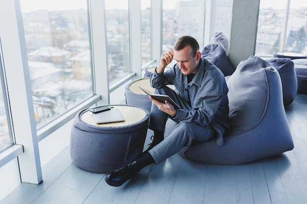 A male businessman in a casual suit resting in a comfortable pouf while working an attractive freelancer in a suit works remotely and checks notes in a notebook