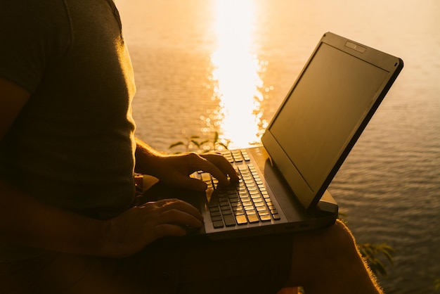 Male businessman with a laptop typing with his hands near the wonderful sunset at the river A man works on a laptop on the background of sunset