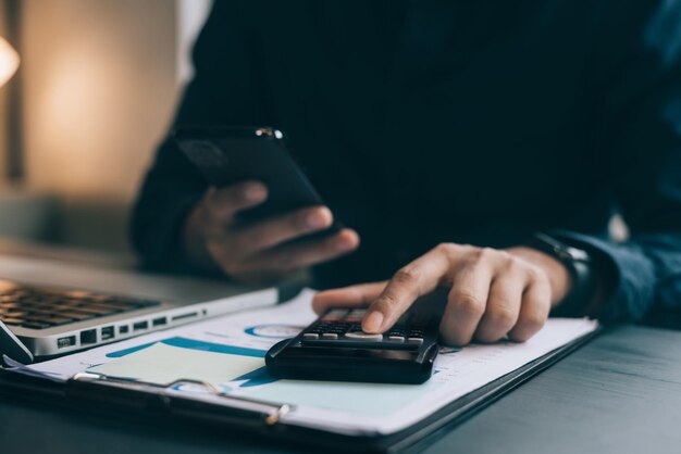 Male businessman working on desk office with using a calculator\
to calculate the numbers and business report talk smart phone on\
office desk finance accounting concept