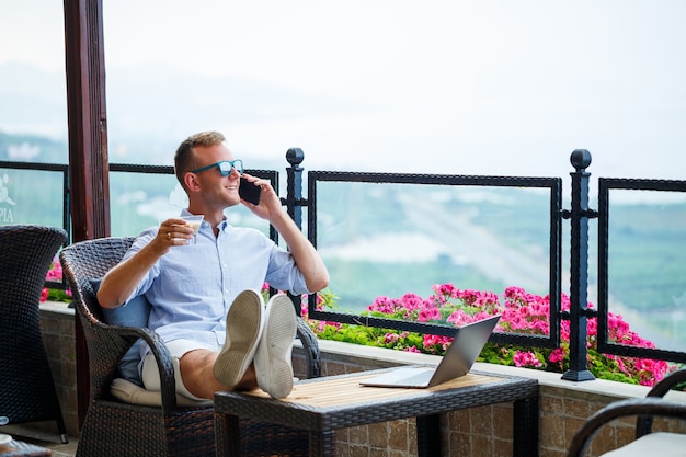 Male businessman working on a laptop on vacation with a beautiful panoramic view. Successful manager drinking coffee and talking on the phone with coffee while traveling