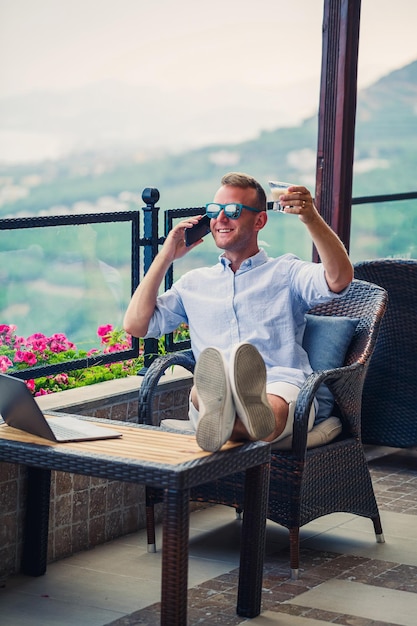 아름다운 탁 트인 전망과 함께 휴가 중 노트북 작업을 하는 남성 사업가 성공적인 매니저는 여행하는 동안 커피를 마시고 커피와 함께 전화 통화를 합니다.