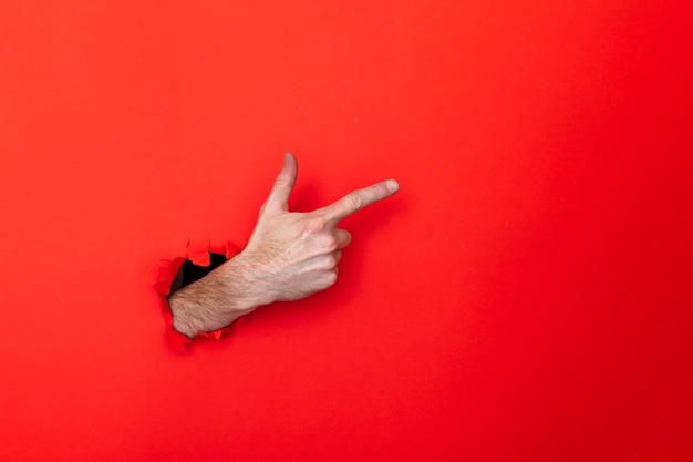 Male caucasian hand through a hole pointing against a red background