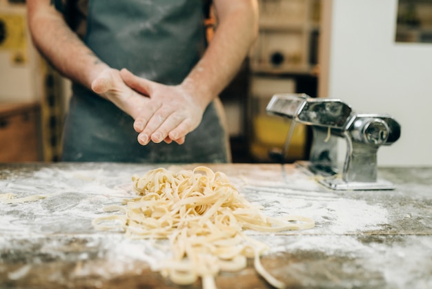 Male chef against wooden kitchen table with uncooked homemade fettuccine and pasta machine. Traditional italian cuisine. Preparation process