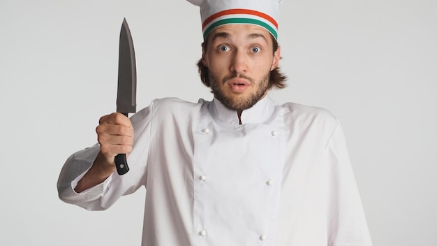 Мужчина-шеф-повар, одетый в белую форму, держит нож, испуганно смотрит в камеру на белом фоне новичок на кухне