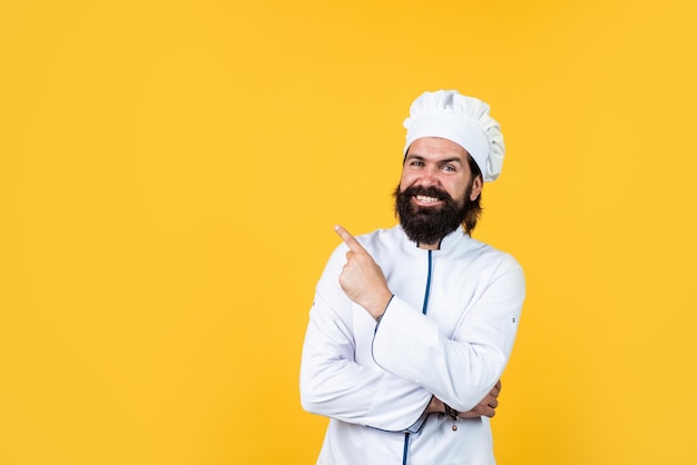 Male chef in hat with beard and moustache on yellow background pointing finger copy space