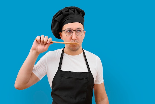 A male chef is tasting food from a spoon on a blue background