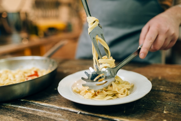 Male chef puts fresh pasta in a plate, pan on wooden kitchen table. Homemade fettuccine preparation process. Traditional italian cuisine