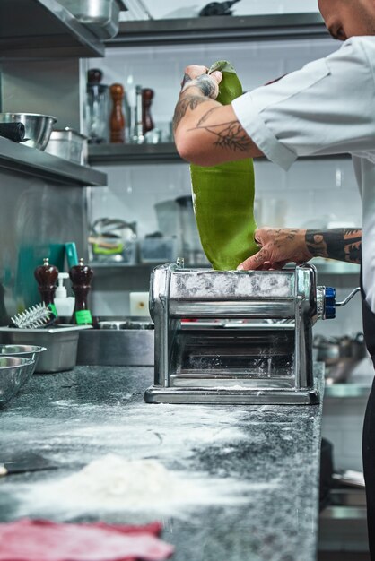 male chef rolling a green dough through pasta machine. cooking process