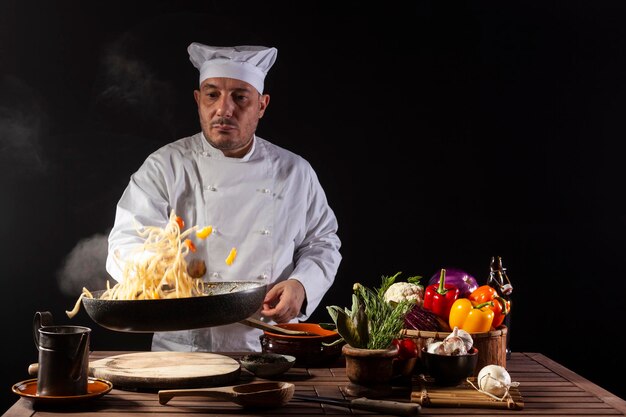 Cuoco unico maschio in uniforme bianca che tiene una padella che salta gli spaghetti con le verdure fresche che volano nell'aria