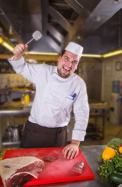 Male chef with a terrible face beats a piece of raw meat. Tenderizing meat. Angry bearded chef cook holding meat mallet.