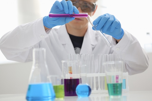 A male chemist holds test tube of glass in his hand overflows a liquid solution of potassium permanganate conducts an analysis reaction takes various versions of reagents using chemical manufacturing.