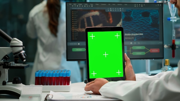 Male chemist scientist holding green mock-up screen tablet sitting at desk. In background technology research, development laboratory with specialist doctor working in high tech design