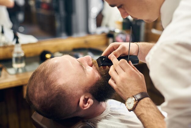 Мужчина-клиент с бородой сидит в парикмахерском кресле Серьезный мужчина с длинной каштановой бородой Современный популярный стиль лесоруба