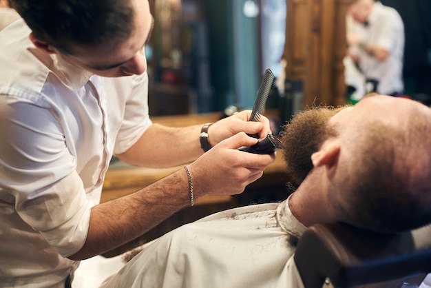 Мужчина-клиент с бородой сидит в парикмахерском кресле Серьезный мужчина с длинной каштановой бородой Современный популярный стиль лесоруба