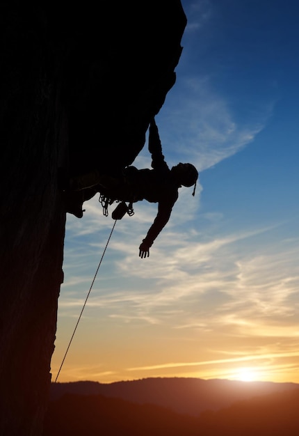 Male climber silhouette hanging on rock one hand down Risky activity in mountains at sunset on cloudy sky Copy space