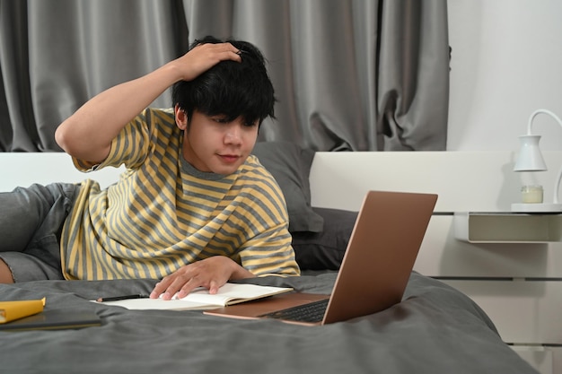 Male college student lying on bed and working with laptop computer