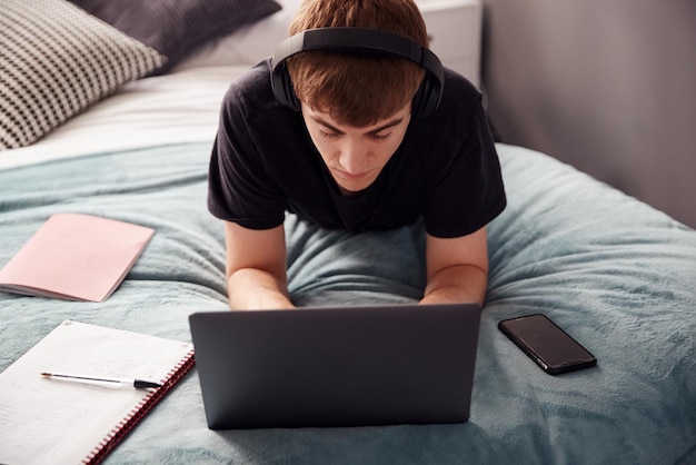 Male College Student Wearing Headphones Lies On Bed In Shared House Working On Laptop