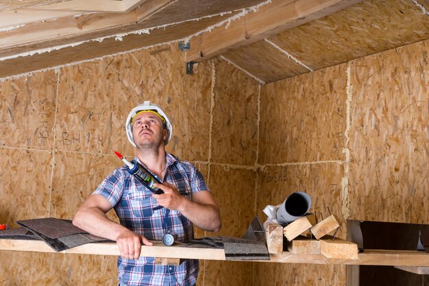 コーキングガンを保持し、露出した粒子合板で未完成の家の中の天井を見上げる男性の建設労働者ビルダー
