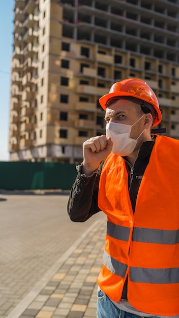 オーバーオールの男性建設労働者と建設中の家の背景に咳をする医療マスク感染の脅威の概念