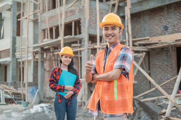 male contractor stands with crossed hands smiling at the camera wearing a safety helmet against a female construction worker in the background