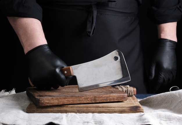 黒のユニフォームと黒のラテックス手袋の男性料理人がまな板の上に大きな鋭い肉ナイフを持っています