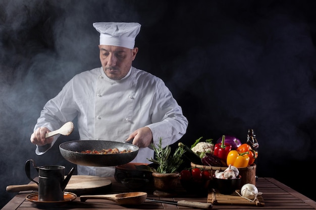 Male cook in white uniform and hat with ladle mixes the ingredients onto the cooking pan before serving while working in a restaurant kitchen