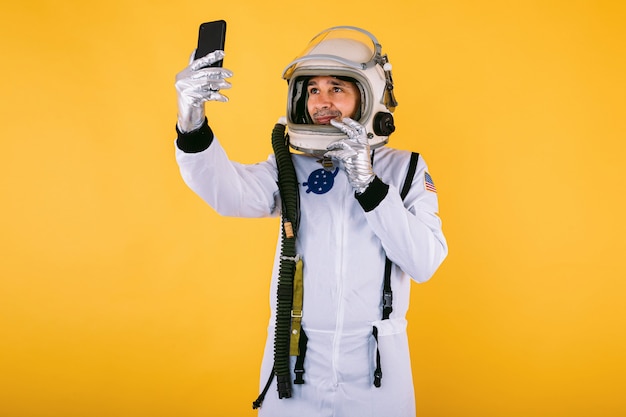 Мужчина-космонавт в скафандре и шлеме, делающий селфи с мобильным телефоном, на желтой стене.