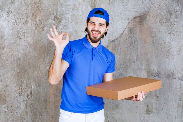 Corriere maschio in uniforme blu che trasporta una scatola di cartone da asporto e mostra un segno positivo con la mano.