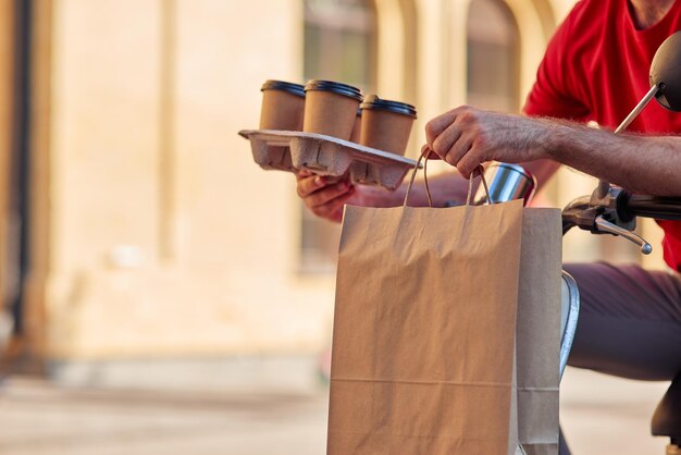 Male courier delivering four coffee cups and paper bag with food by scooter