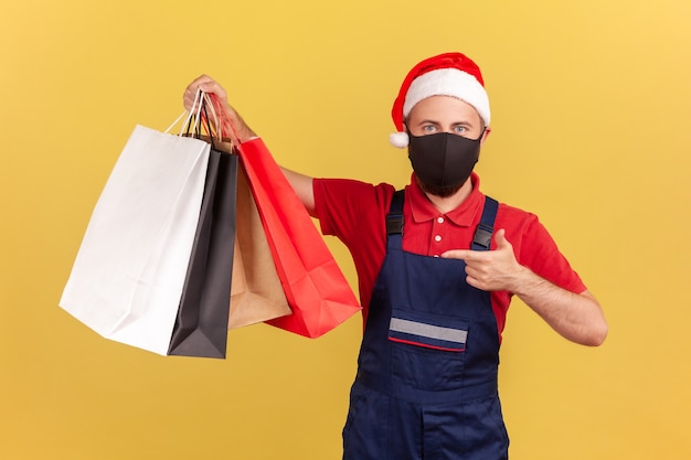 Male courier in mask and holiday santa claus hat pointing finger at shopping bags