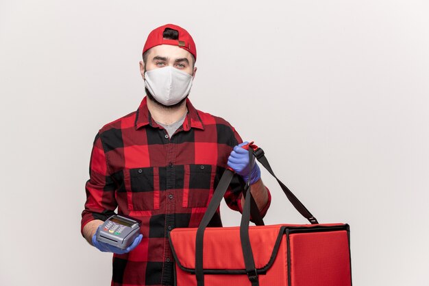 Male courier in red cap, flannel, protective mask and gloves carrying bag with orders and payment machine during work in quarantine period