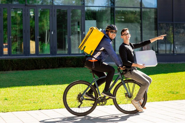 Male courier with thermal bag is carrying a guy with a box on a bicycle trunk. 