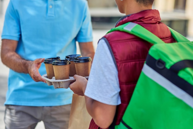 Male courier with thermo bag delivering four coffee cups to customer while standing outdoors