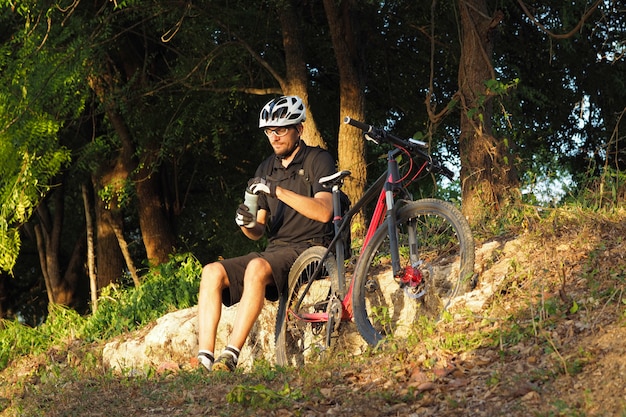 Male Cyclist Sitting on Rocks and Drinking Water in Forest.
