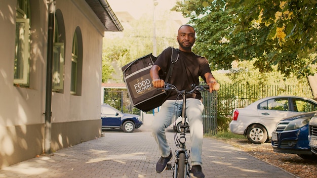 Male delivery courier riding bike with thermal backpack to give fastfood order package to clients. Going to front door entrance to deliver takeaway meal, restaurant delivery service.
