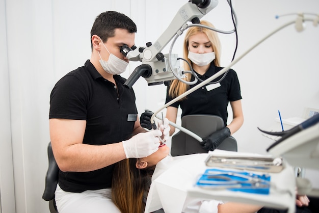 Male dentist and female assistant treating patient teeth with dental tools - microscope, mirror and drill at dental clinic office. Medicine, dentistry and health care concept.