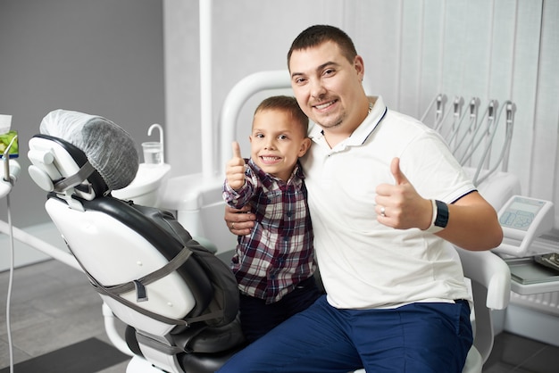 Male dentist and his young child client are sitting together hugging and showing thumbs up in a dental office, both being satisfied and happy after the dental treatment.