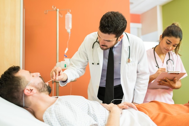 Male doctor checking throat of patient while female healthcare\
worker making reports at hospital